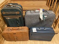 Four suitcases grouped together on wooden floor below wooden staircase: dark green canvas with brown leather, dark gray leather, tan crocodile pattern leather, navy blue leather