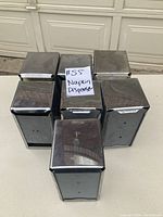 Seven rectangular stainless steel napkin dispensers placed in two rows on a concrete floor indoor, showing front and top surfaces reflecting surrounding light.