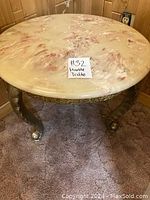 Photo of a round coffee table with a marble top and ornate metal legs, positioned on a carpeted floor against wood-paneled walls.