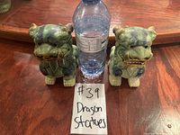 Pair of small green porcelain dragon statues placed beside a water bottle for scale on a wooden surface.