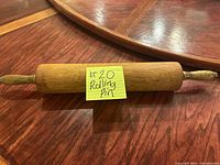 Photo of a wooden rolling pin on a wooden surface with a yellow note labeled '#20 Rolling Pin'. The pin shows signs of use and has handles on both ends.