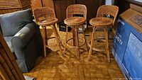 Three wicker swivel bar stools grouped together on a tile floor next to a grey couch and boxes.
