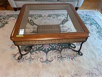 Full overview of coffee table showing square wood frame with inset glass top, metal frame legs with curled details, and woven lower shelf.