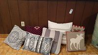 Seven cushions arranged on a floor against wood paneling showing diverse patterns and colors including moose motif, stripes, checkered, and solid white.
