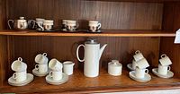 Two espresso sets on wooden shelf showing coffee pot, sugar bowl, creamer, demitasses, saucers, and metallic cup holders.