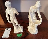 Two white figurines: one male standing nude with sling over shoulder on rectangular base, one female leaning on pedestal holding a bowl, seen from front