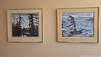 Two framed prints on beige wall featuring Canadian landscape scenes by Group of Seven artists.