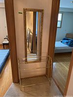 Wide angle of the mirror mounted on the wall above the metal blanket stand showing full view of both items and their gold-toned finish.