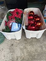 Two white storage bins placed on the ground filled with Christmas decorations including red ornaments, ribbon, faux poinsettia, and garland. Photo to show overall lot contents.