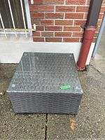 Full front view of square patio table with glass top showing grey woven resin wicker base on concrete outdoor surface.