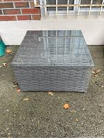 Front view of square resin wicker patio table with glass top on outdoor concrete surface.