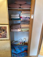 Shelves with folded towels in various colors and three mauve accent cushions on top shelf, bag of William Ashley twin bed sheets and folded Thomas the Tank Engine twin comforter at bottom.