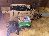 Photo showing two black wired Xbox controllers, power supply cables, and five Xbox game cases stacked inside a cardboard box on the floor.