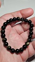 Close-up image showing the bracelet being held, displaying round polished golden sheen obsidian beads with visible golden flashes among the black beads.