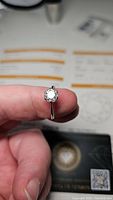 Moissanite ring held between fingers showing the round brilliant cut gemstone and polished silver band with certification papers blurred in background.