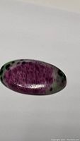 Oval polished ruby zoisite cabochon with prominent dark red ruby areas, green zoisite background and some black spots, shown on white background.