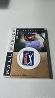 Photo of an Adam Hadwin golf ball marker card with PGA Tour logo ball marker attached, facing upwards showing the front design of the card and marker.