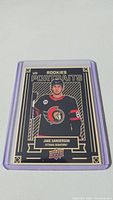 Photo shows Jake Sanderson rookie hockey card in protective case, featuring him in Ottawa Senators uniform with UD Rookies Portraits branding.