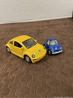 Yellow 1998 New Beetle diecast car alongside smaller blue classic Beetle model on brown surface