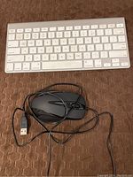 Apple wired keyboard with white low-profile keys showing wear and a black wired mouse with USB cord, placed on a textured brown surface.