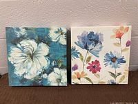 Lot showing both floral art canvases side by side, one with large white flowers on teal background and the other with multicolor flowers on white background