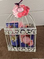White decorative bird cage with floral patterns filled with various Casa Belle bath and body products, viewed from the front showing labeled bath cream and box.