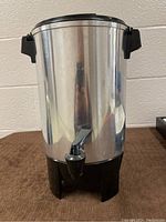 Front view of stainless steel coffee urn with black handles and spigot on brown surface.
