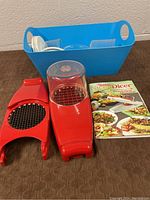 Full view of Genius Nicer Dicer chopper with main body, dome lid, red cutting grid insert, additional slicing grid, blue storage tub with cups and white parts, and instruction manual.