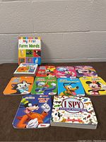 Photo of all eleven kids board books arranged flat on a brown surface against a gray wall, showing colorful covers featuring Disney characters and educational themes.
