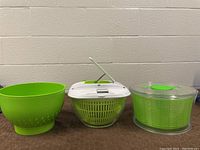 Three kitchen items side by side on table: a lime green plastic colander, a salad spinner with clear bowl and white lid, and a vegetable chopper with clear container and green basket