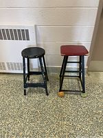 Two metal stools, one round black with chipped paint and one square red with various colored protective feet.