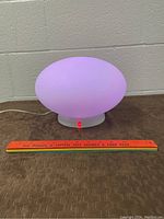 Oval-shaped mood lamp on with soft lavender light, cord attached, ruler in front for scale.