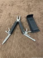 Mountain Quest multi tool unfolded showing pliers, knife, saw, and other tools next to the black nylon carrying case.