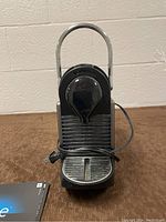 Front view of the Nespresso Pixie espresso machine showing black and silver build with cord wrapped around the base and drip tray visible.