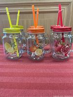 Front view of six glass mason jar mugs arranged in two rows showing colorful fruit print designs, matching lids, and matching reusable plastic straws.