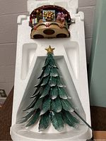 Musical Christmas tree candle holder in white styrofoam packaging, metal painted green with textured layered leaves, snow accents, and gold star on top.