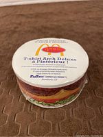 Close-up of sealed white round compressed packaging with McDonald's Arch Deluxe logo and text in English and French indicating T-shirt inside.