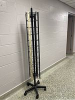 Full view of the over five feet tall black metal retail display rack against a wall, showing the vertical grid shelves and five-legged wheeled base.