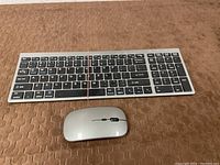 Wireless keyboard and wireless mouse on a brown textured surface, keyboard has black keys with white and blue lettering, mouse is silver with scroll wheel.