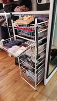 Full view of the rack showing six white wire baskets filled with folded textiles and accessories, positioned in a closet-style space on hardwood flooring.