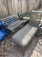 Two-seat outdoor bench next to a patio lounge chair on a brick patio against a wooden fence, showing weathered wooden slats and cast iron armrests on the bench along with mesh sofa lounge chair.