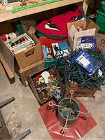 Overview of box and containers filled with Christmas lights, ornaments, tree skirt and other decorations on floor.