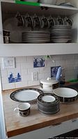 Multiple Mikasa Intaglio dinnerware items including plates, bowls, cups, a covered casserole dish, and platters on kitchen counter and shelf.