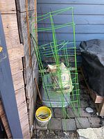 Photo showing multiple green wire tomato cages and plant stakes leaning against wooden wall, with bag of soil or plant feed and galvanized bucket below.