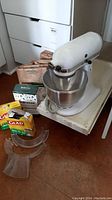 Front view of white KitchenAid mixer with bowl attached, two plastic shields and three packs of paper organic waste bags on white platform.