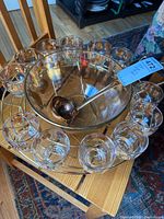 Overview of punch bowl, ladle, metal stand, and 12 glass cups on and around wooden chair.