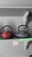 Two cast iron teapots on a shelf, one red and one purple, showing their size and design