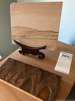 Two original tile art pieces placed on wood surface with one on wooden stand showcasing layered sediment patterns and carved abstract figures.