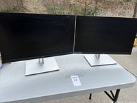 Two HP L27m monitors side by side on a folding table showing front view with black screens and silver stands