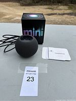 Apple HomePod mini speaker with original box, power cable, and instruction manual laid out on table.